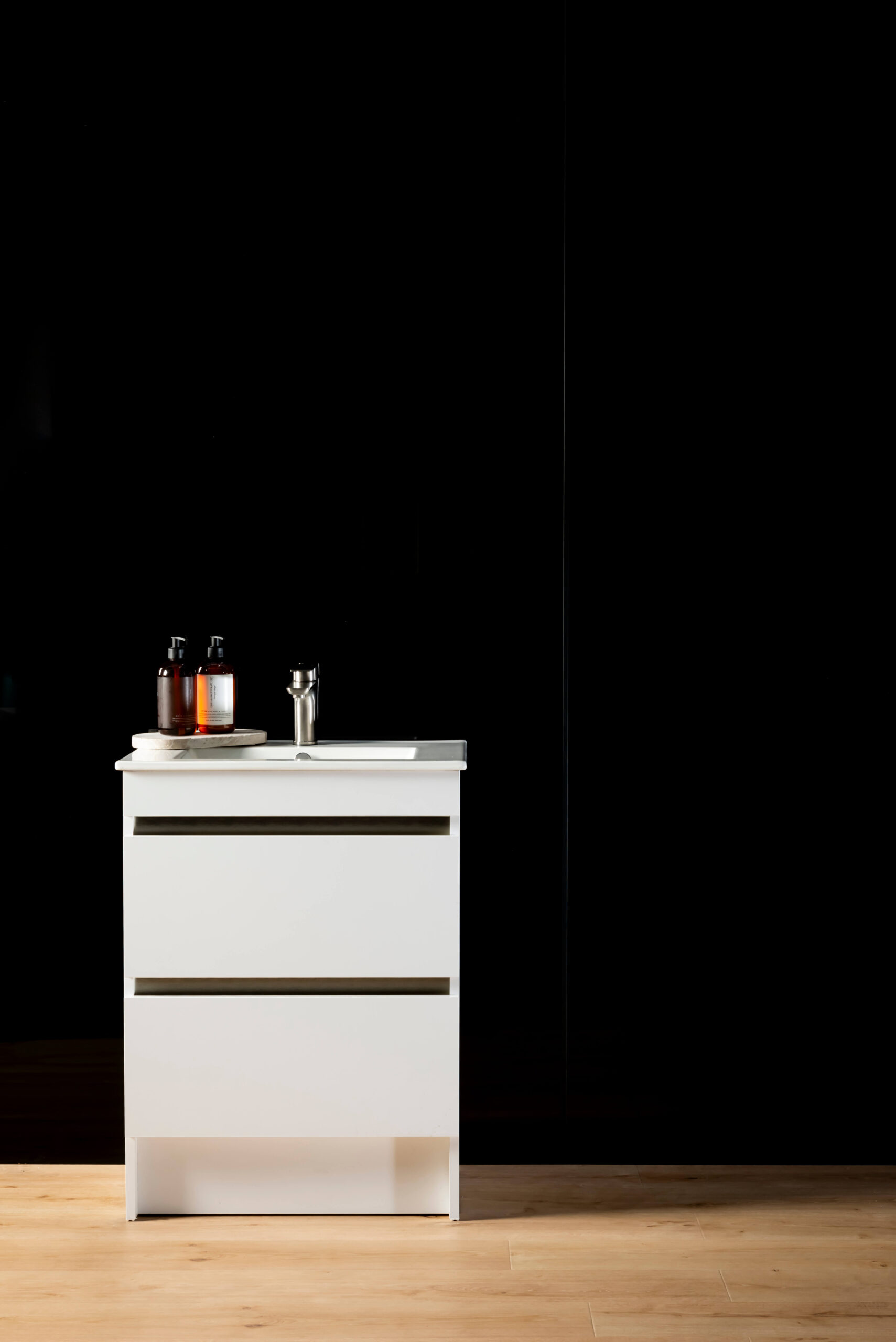 A black panel wall joined with a black joiner. A white vanity in front with soap and a wooden floor in the forefront.