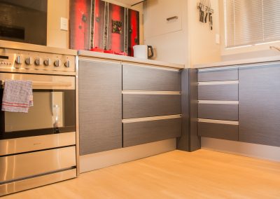 A modern kitchen with stainless steel appliances, blue/grey cabinetry, and light wood flooring. The cabinets have sleek silver handles, and matching silver wet wall toe kicks. Red and black artwork is displayed above the counter